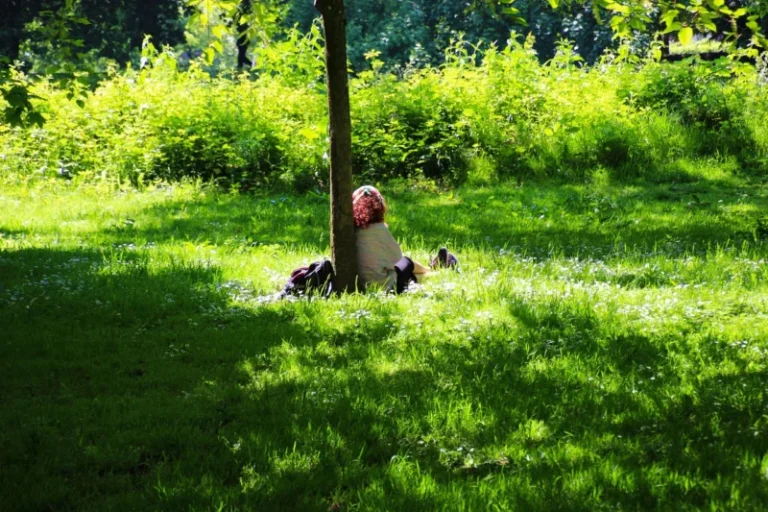Mlada žena tiho sedi na travi u zelenom parku, izgleda smireno i zaokupljeno sobom u meditaciji
