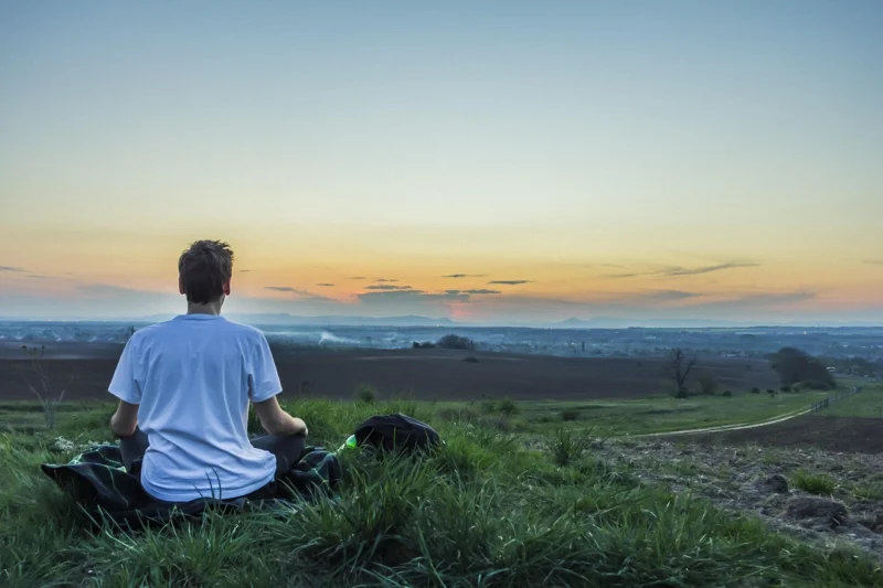 Osoba sedi u pozi lotos dok meditira u prirodnom okruženju pri zalasku sunca, okružena mirom i tišinom
