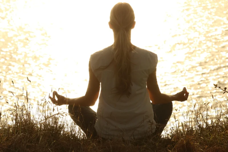 Žena meditira u prirodi tokom zalaska sunca, sedeći u joga pozi na travi, okružena spokojem i tišinom