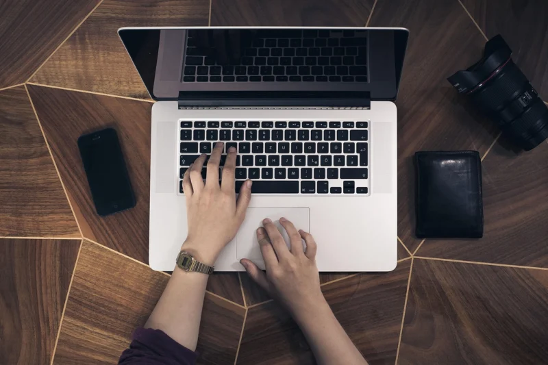 Ruke tipkaju na tastaturi laptopa, dok je mobilni telefon pored njih na stolu