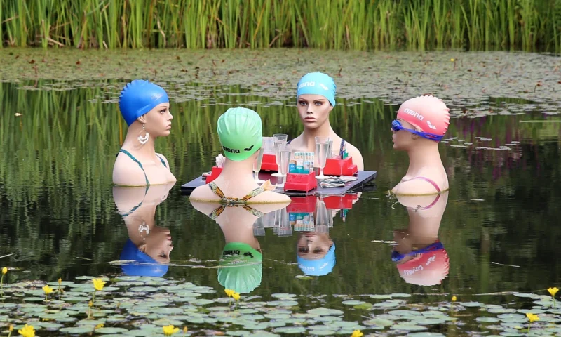 Četiri žene sa kapama za plivanje sede ili stoje pored plitke vode u umetničkoj instalaciji osvetljenoj prirodnim svetlom u botaničkoj bašti, dok razgovaraju i reflektuju svoje misli.