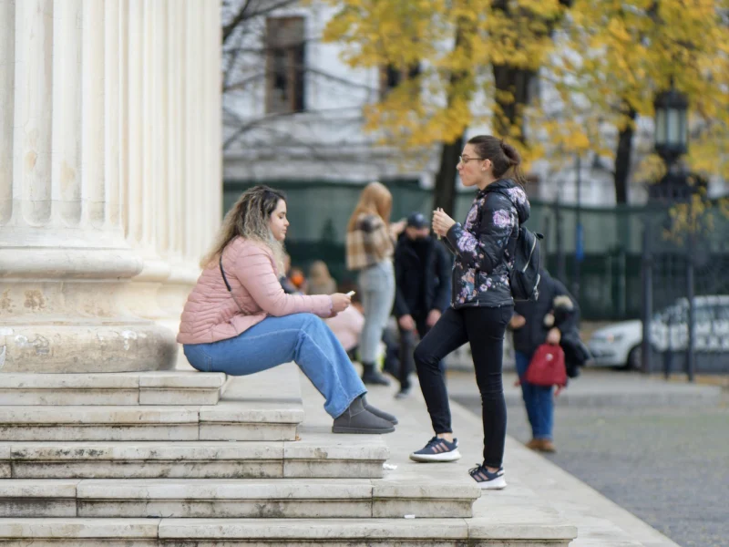 Dve mlade devojke razgovaraju i sede ispred zgrade sa stubovima na otvorenom prostoru