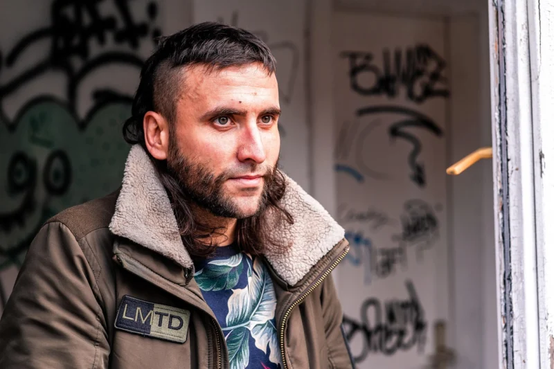 Profil mladog muškarca sa bradom koji zamišljeno gleda kroz prozor dok mu se u pozadini vide grafiti na zidu
