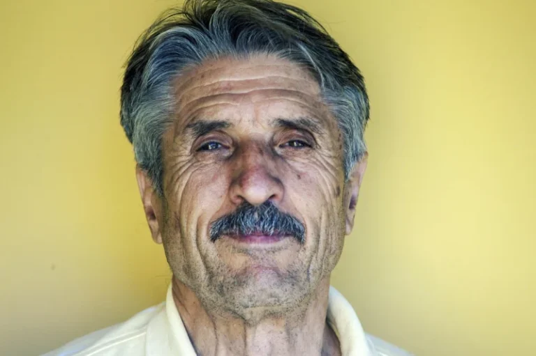 Stariji čovek sa dugom sedom kosom i brkovima, koji se smireno osmehuje gledajući pravo u kameru
