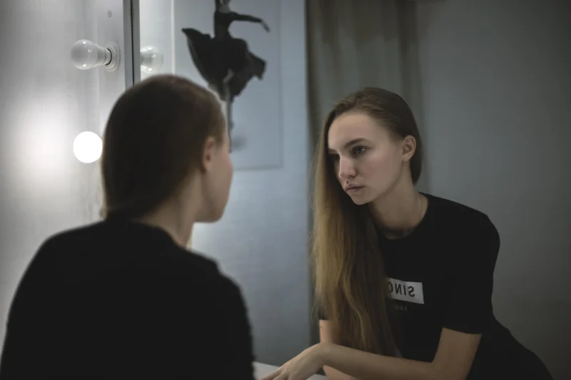 Žena stoji ispred ogledala u kupatilu i zamišljeno posmatra svoj odraz