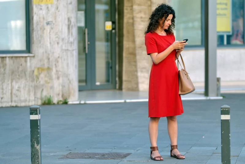 Mlada žena u crvenoj haljini stoji na trotoaru u urbanom okruženju, držeći telefon i torbu, dok pažljivo gleda u ekran svog pametnog telefona.
